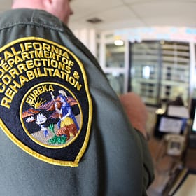 Close-up of a California Department of Corrections and Rehabilitation uniform patch on a green shirt, featuring the word "Eureka" and an emblem depicting individuals in a historical scene. The background shows a blurred office setting.