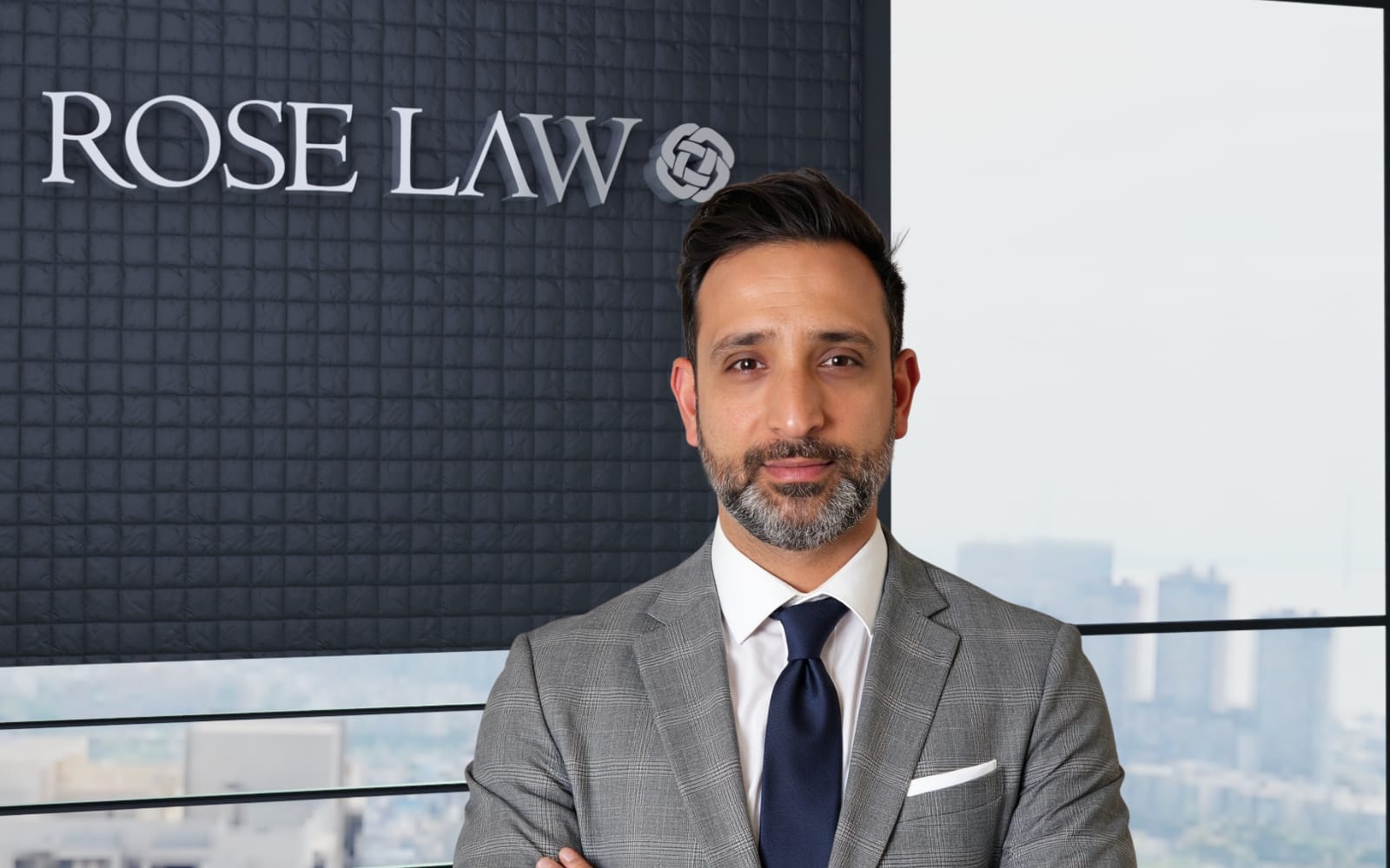 Mehran Tahoori, wearing a gray suit and blue tie, stands with arms crossed in a modern office setting. A logo reading "ROSE LAW" is visible in the background, alongside a city skyline seen through large windows.