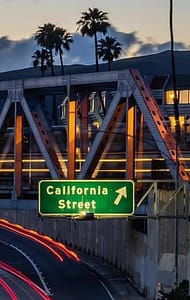 A green directional road sign for "California Street" is visible on a bridge, surrounded by palm trees and illuminated by nearby lights. The sky is dusky with a gradient of blue and orange, highlighting a busy highway below.