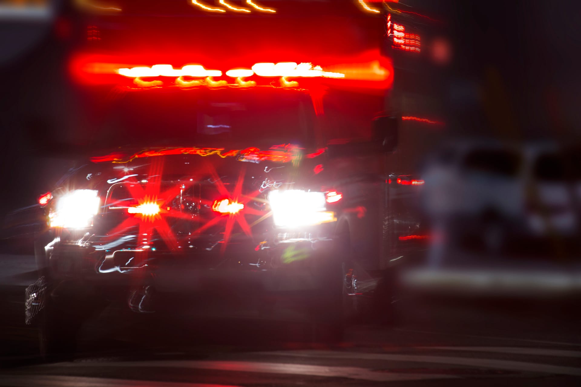 A motion-blurred image of an ambulance with flashing red lights on a city street at night.