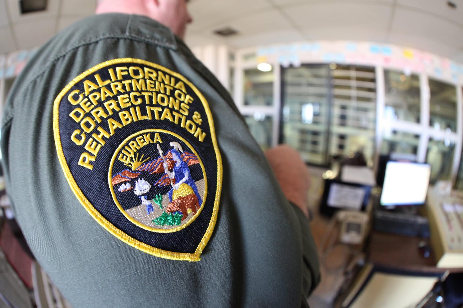 Close-up of a California Department of Corrections and Rehabilitation uniform patch on a green shirt, featuring the word "Eureka" and an emblem depicting individuals in a historical scene. The background shows a blurred office setting.