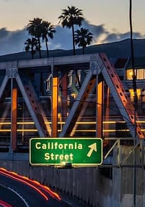A green highway sign indicating "California Street" is illuminated at night. The sign is near an overpass with metal trusses reflected in the lights. Palm trees are visible against a twilight sky, adding to the California ambiance.