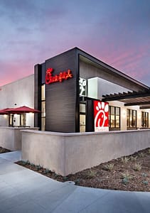 Exterior view of a Chick-fil-A restaurant featuring a modern design. The building has large windows, a prominent illuminated logo, and patio seating under red umbrellas, with landscaping visible in the foreground.