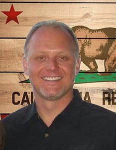 Labor and Employment Lawyer Joe Rose wearing a black open collar shirt standing against a wood California flag