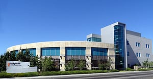 A modern medical center building with a curved stone façade and large glass windows, set against a clear blue sky. The entrance features a sign with 'Dignity Health' prominently displayed. Nearby, trees and greenery are visible.