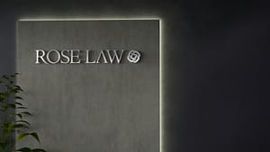 A modern reception area featuring a wall sign that reads "ROSE LAW" in sleek font, illuminated by soft lighting. A small green plant is visible in the lower left corner against a textured gray wall.