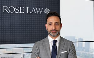 Mehran Tahoori, wearing a gray suit and blue tie, stands with arms crossed in a modern office setting. A logo reading "ROSE LAW" is visible in the background, alongside a city skyline seen through large windows.