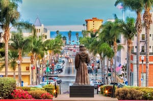 A view of a street lined with palm trees leading to the ocean, with a statue of a robed figure in the foreground. Buildings and vehicles are visible, and the sky shows a gradient from blue to light gray.