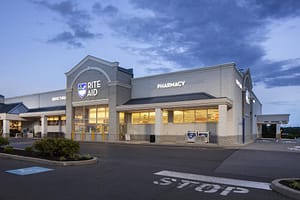 Exterior view of a Rite Aid pharmacy building during twilight, showcasing large windows with illuminated signage. The parking lot is visible in front of the store, marked with a stop sign and landscaping along the edge.