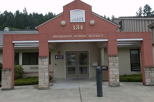 Exterior view of the Bremerton School District building, featuring a prominent red entrance with a triangular roof. The building displays the number "134" and the name "Bremerton School District" above the main door, surrounded by landscaped greenery.