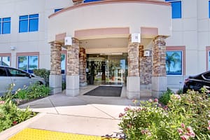 The entrance to Rose Law APC in Ventura features a glass door framed by stone pillars. Flanking the door are two vehicles parked on a paved walkway, with landscaped greenery and colorful flowers in the foreground.