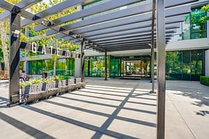 The entrance of a modern building featuring a large sign that reads "GATE ONE." A covered walkway with metal beams extends overhead. Planters and greenery are visible, providing a natural contrast to the building's glass facade.