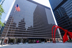 A large modern building with a glass facade reflects the surrounding architecture, featuring an American flag on the left. In the foreground, a large red sculpture adds contrast to the neutral tones of the building. This image relates to discussions about national origin and ethnicity discrimination in employment.