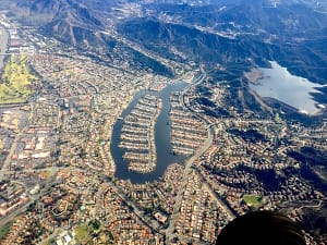 Aerial view of a residential area featuring a large marina with numerous boats docked, surrounded by houses and hills. The landscape includes a nearby lake and a blend of urban and natural settings, showcasing the connection between the community and the waterfront.