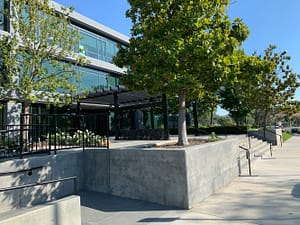 A modern office building with large glass windows surrounded by trees and landscaped areas. The exterior features a concrete patio with planters, accessible ramps, and walkways leading to the main entrance.